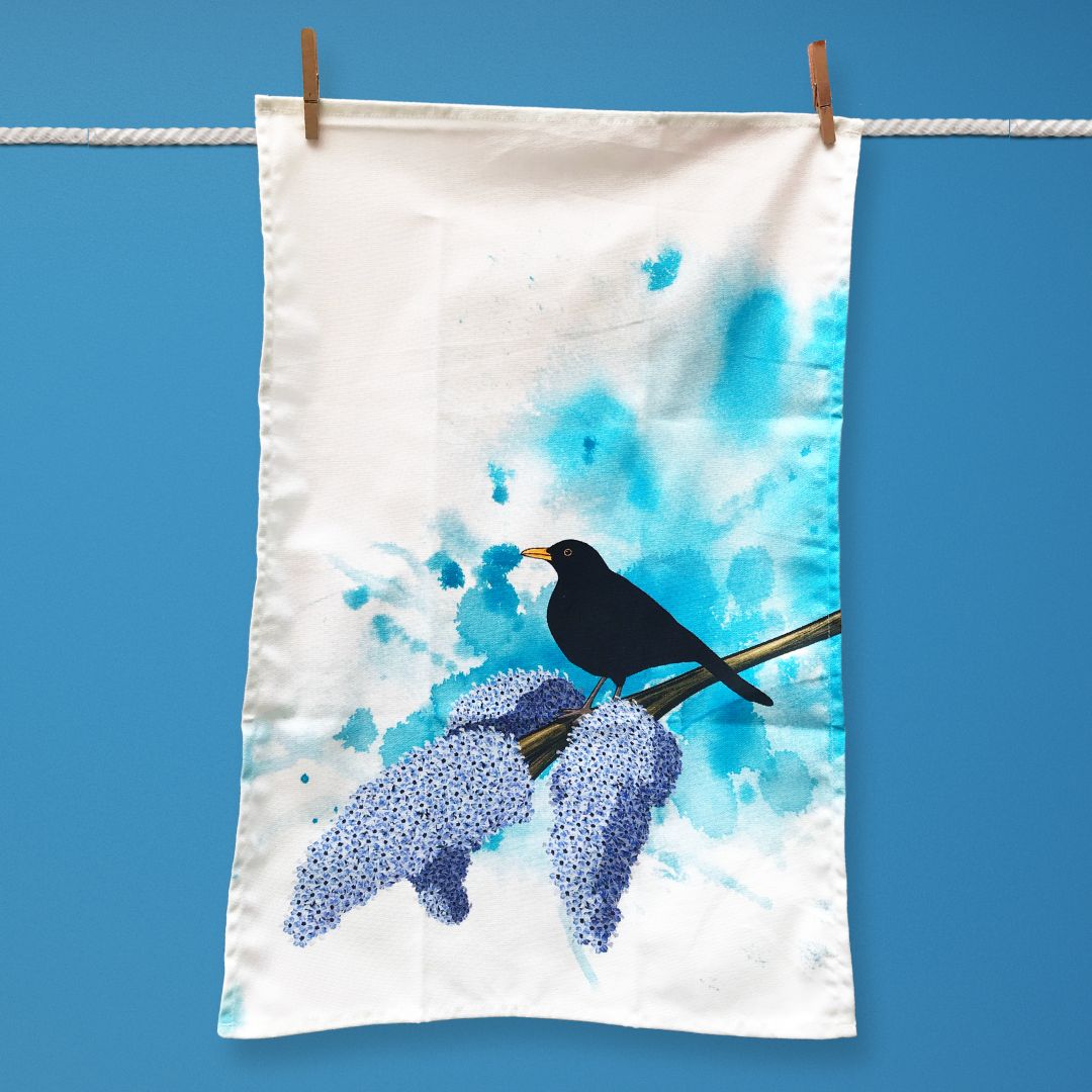 A tea towel hanging on a washing line. The illustration on the tea towel shows a male black bird perched on a branch of lilac with three full purple blooms on the end. The background is an inky wash in bright turquoise. 