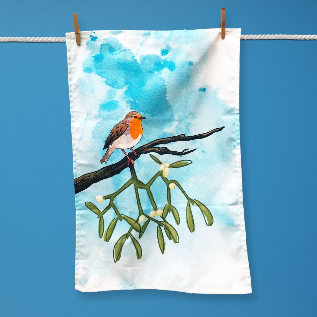 A tea towel hanging on a washing line. The illustration on the tea towel shows a robin perched on a branch of mistletoe. The background is an inky wash in bright turquoise. 