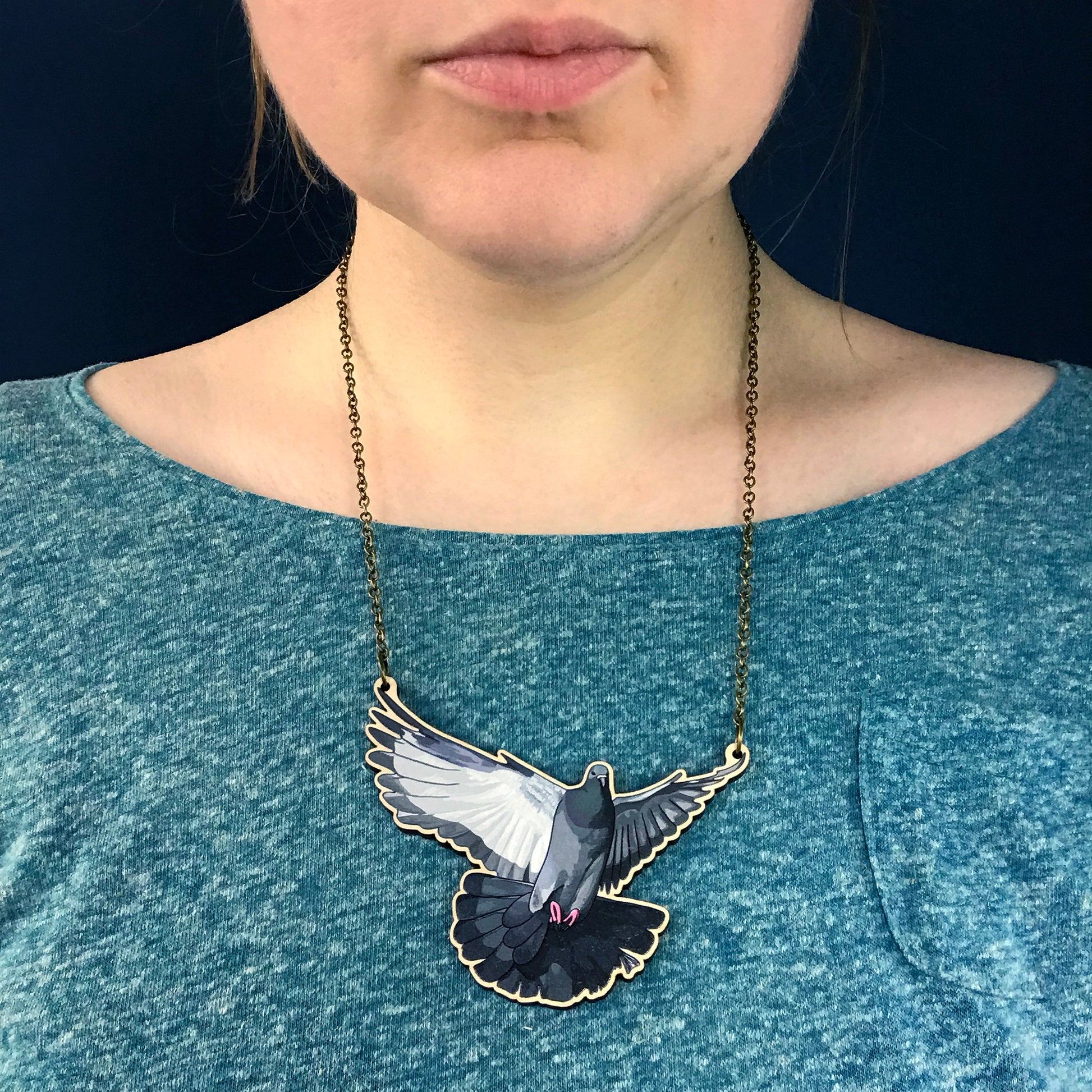 Flying pigeon necklace by Loadofolbobbins. A laser cut wooden necklace that shows an illustrated pigeon with its wings spread out flying. The necklace hangs on an antique bronze chain. It is being worn by me a white woman wearing a light turquoise speckled top. You can just see the bottom of my face and neck.