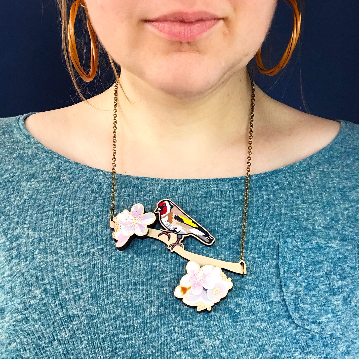 Goldfinch and blossom necklace by Loadofolbobbins. A laser cut wooden necklace that shows an illustrated goldfinch on a tree branch that has two light pink collection of blossoms standing proud. The necklace hangs on an antique bronze chain. It is being worn by me a white woman wearing a light turquoise speckled top. You can just see the bottom of my face and neck.