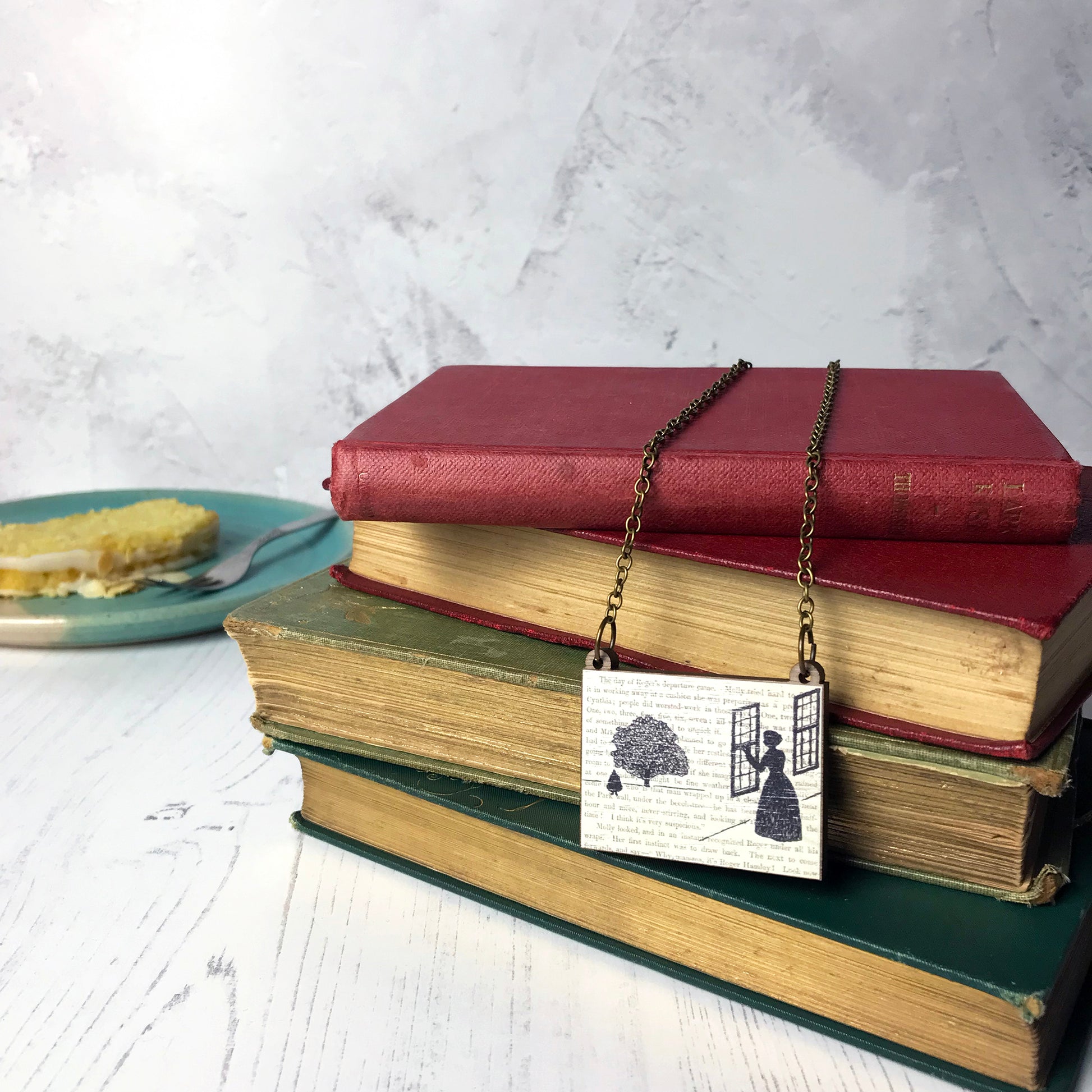 A laser cut wooden necklace by loadofolbobbins showing a silhouette scene inspired by the novel Wives and Daughters by Elizabeth Gaskell. The scene shows Molly looking out of the window at  Roger standing under a large tree. The scene is printed against original text from the novel. The necklace is drapped over and dangling on a pile of old books.