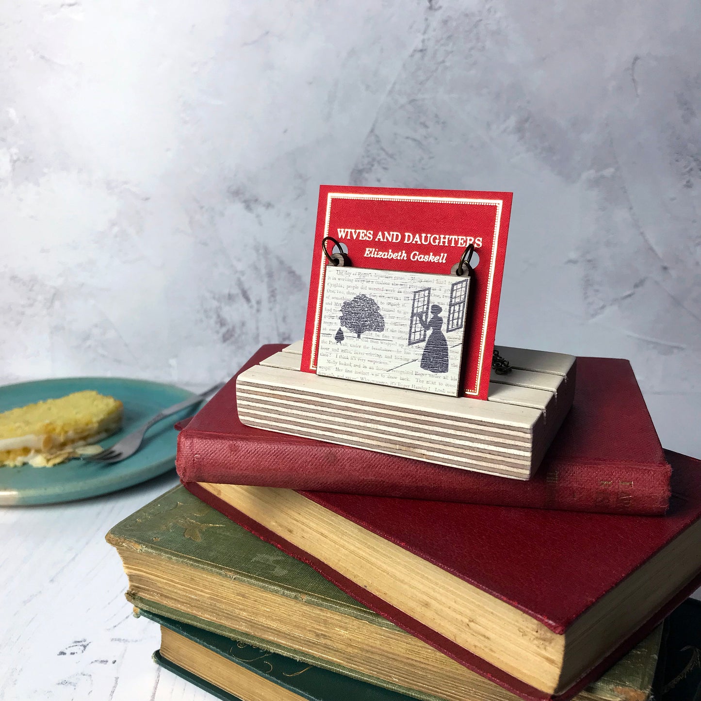 A laser cut wooden necklace by loadofolbobbins showing a silhouette scene inspired by the novel Wives and Daughters by Elizabeth Gaskell. The scene shows Molly looking out of the window at Roger standing under a large tree. The scene is printed against original text from the novel. The necklace is mounted on a red backing card with gold foil lettering detail and made to look like the front of a book and standing on a pile of old books.