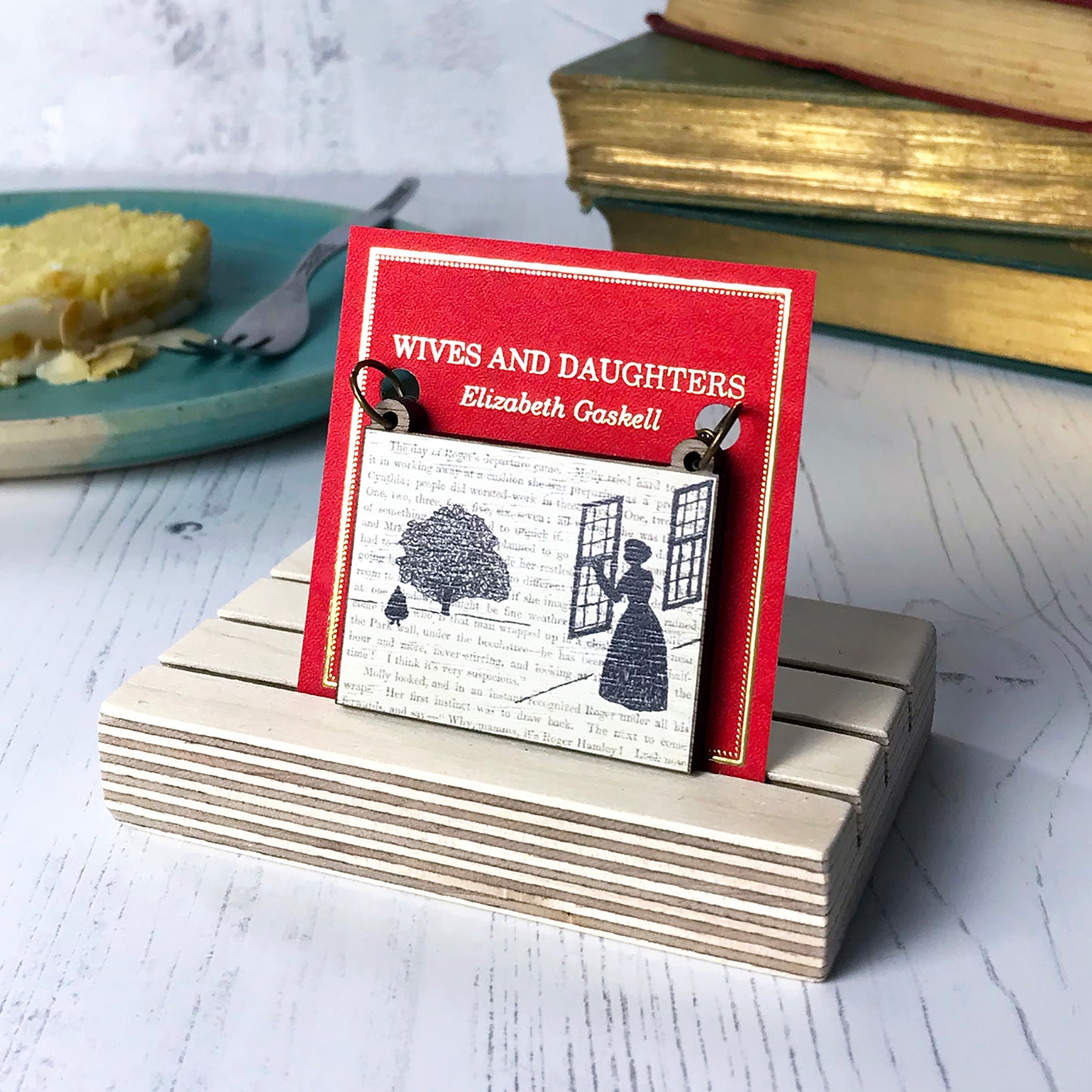 A laser cut wooden necklace by loadofolbobbins showing a silhouette scene inspired by the novel Wives and Daughters by Elizabeth Gaskell. The scene shows Molly looking out of the window at Roger standing under a large tree. The scene is printed against original text from the novel. The necklace is mounted on a red backing card with gold foil lettering detail and made to look like the front of a book.