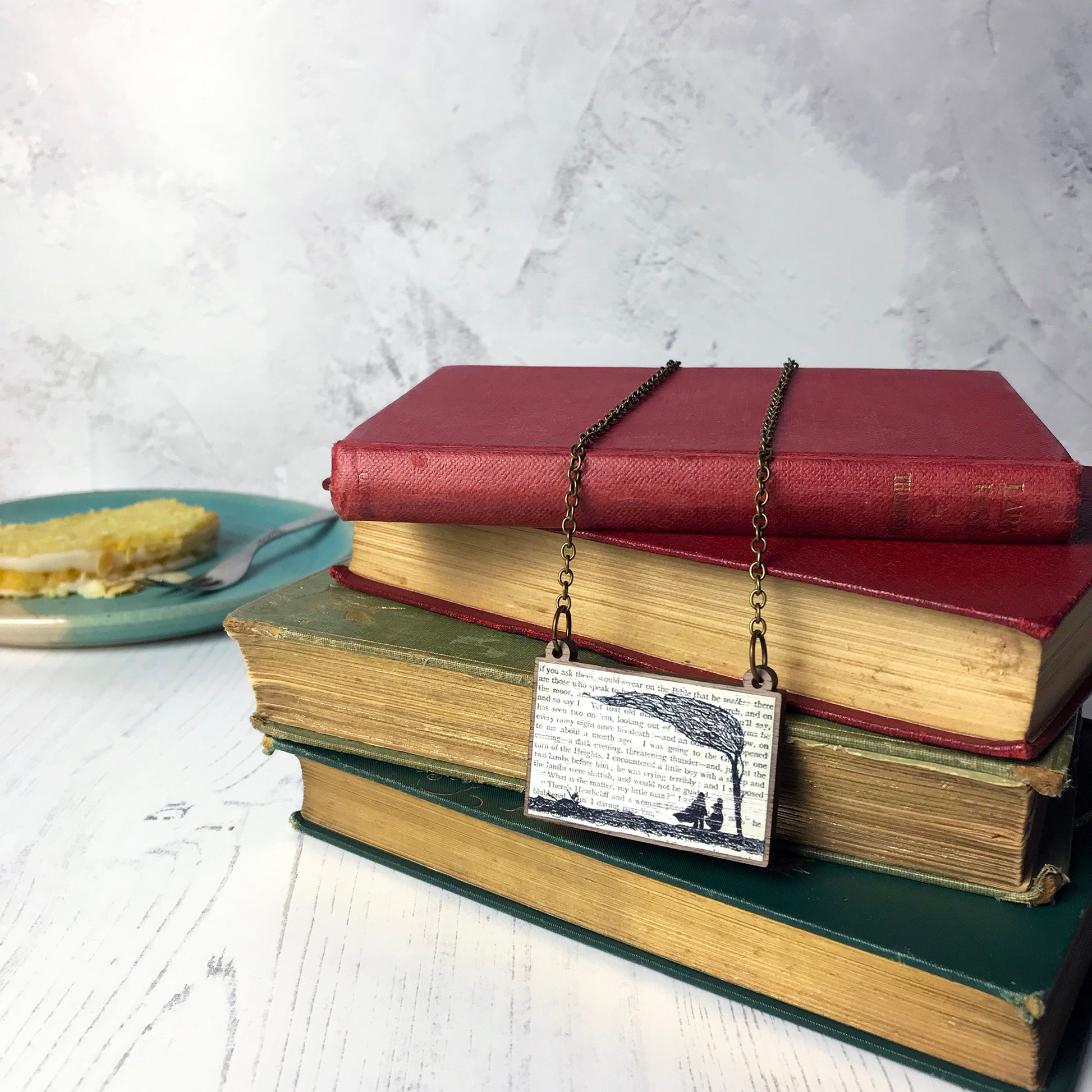 A laser cut wooden necklace by loadofolbobbins showing a silhouette scene inspired by the novel Wives and Daughters by Elizabeth Gaskell. The scene shows Molly looking out of the window at  Roger standing under a large tree. The scene is printed against original text from the novel. The necklace is drapped over and dangling on a pile of old books.